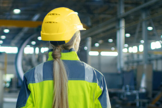 A woman with a ponytail is wearing a helmet and high-vis jacket in an industrial environment.