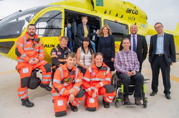 Air ambulance staff (operative and desk-based) pose outside of an air ambulance with a man in a wheelchair.