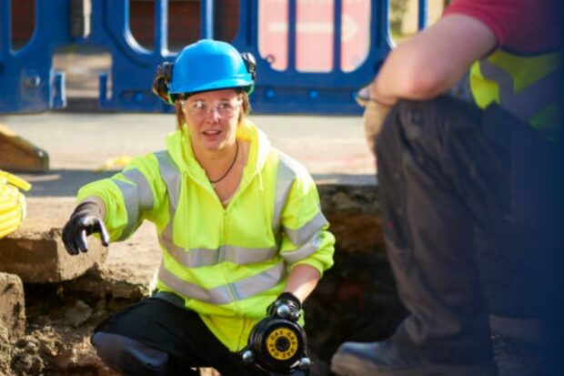A woman, wearing a yellow high-vis jacket and a blue hardhat, squats inside a hole dug in the ground holding equipment.